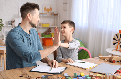 A supportive adult giving a high-five to a smiling young boy during a learning activity, highlighting positive interaction and autism awareness in an educational setting.