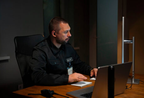 Security guard working at a dimly lit desk, monitoring activity on multiple screens as part of his Prevent Duty responsibilities, focused in a professional environment.